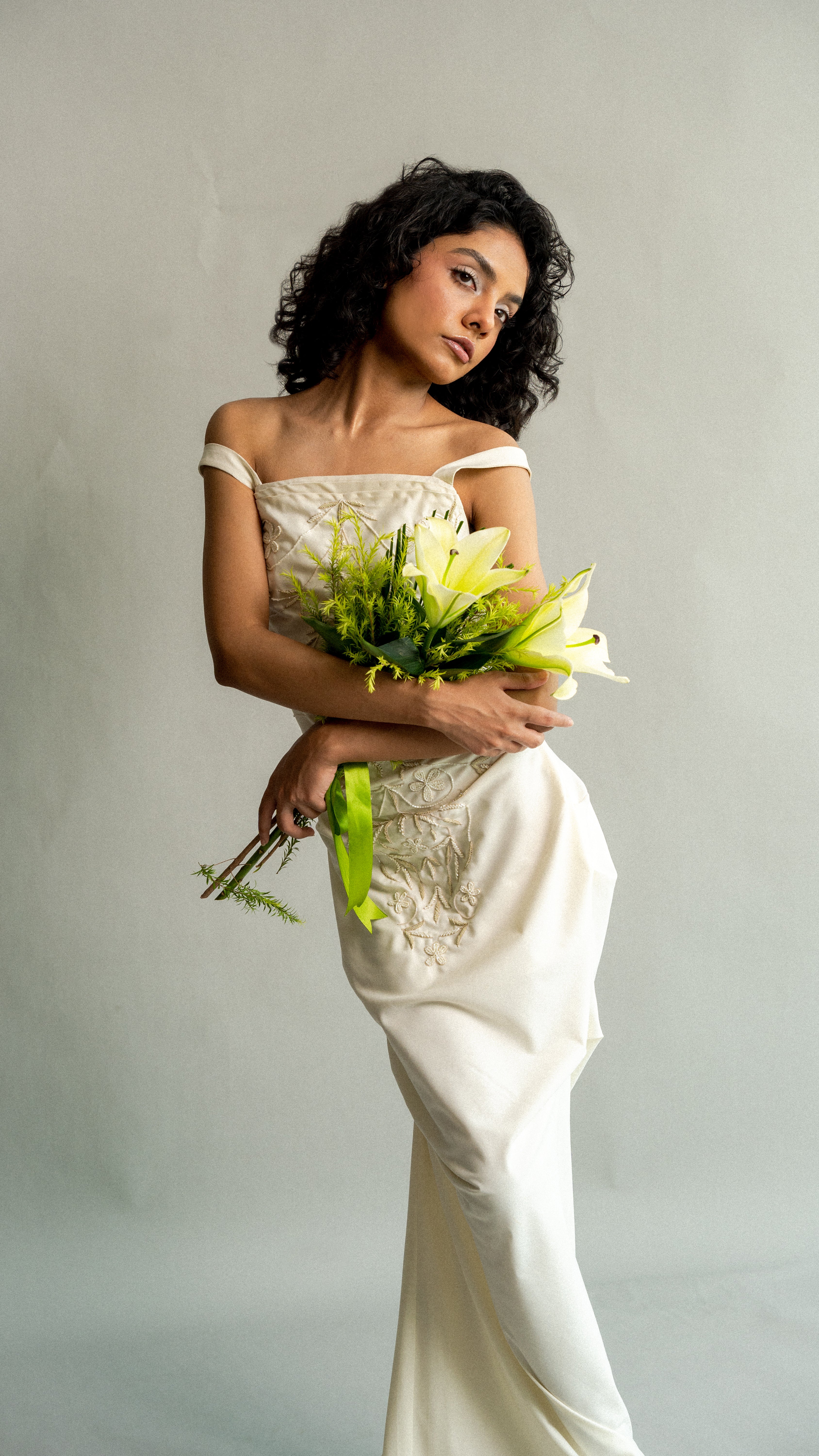 Woman in a white lace dress holding a bouquet of flowers against a plain background