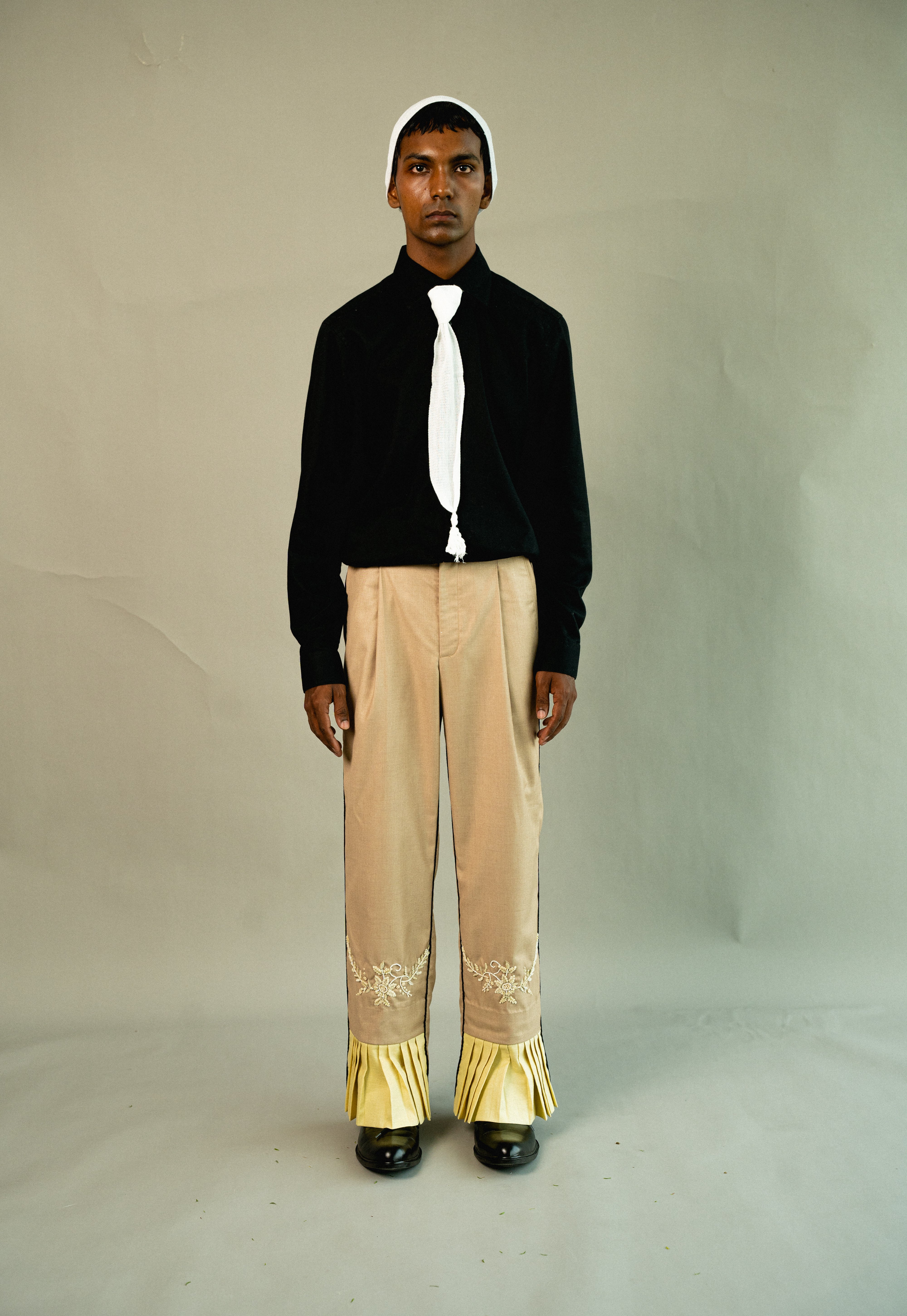Person wearing a black jacket, white shirt, and beige pants with gold accents on a plain background