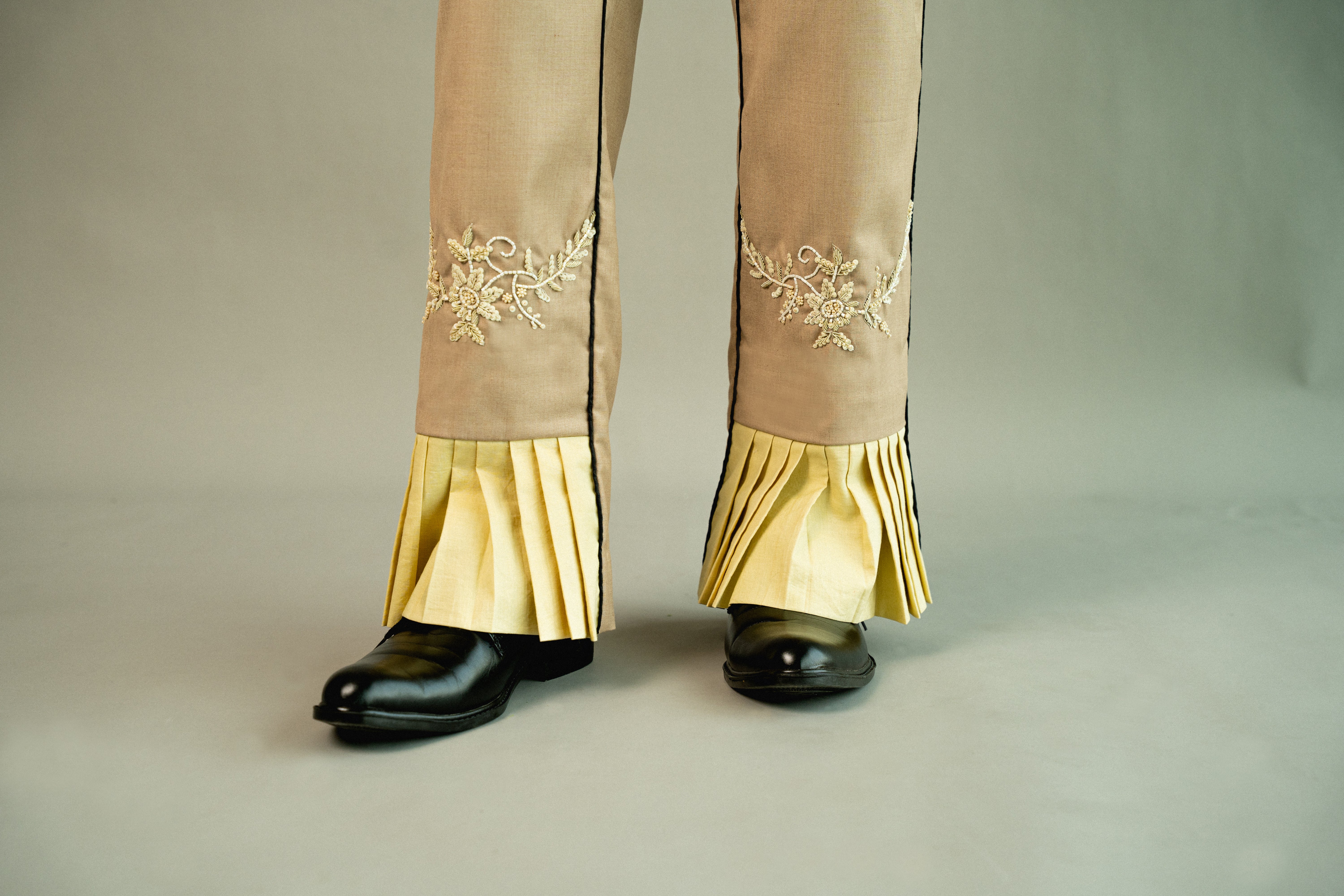 Beige pants with gold embroidery and ruffles worn with black shoes on a neutral background