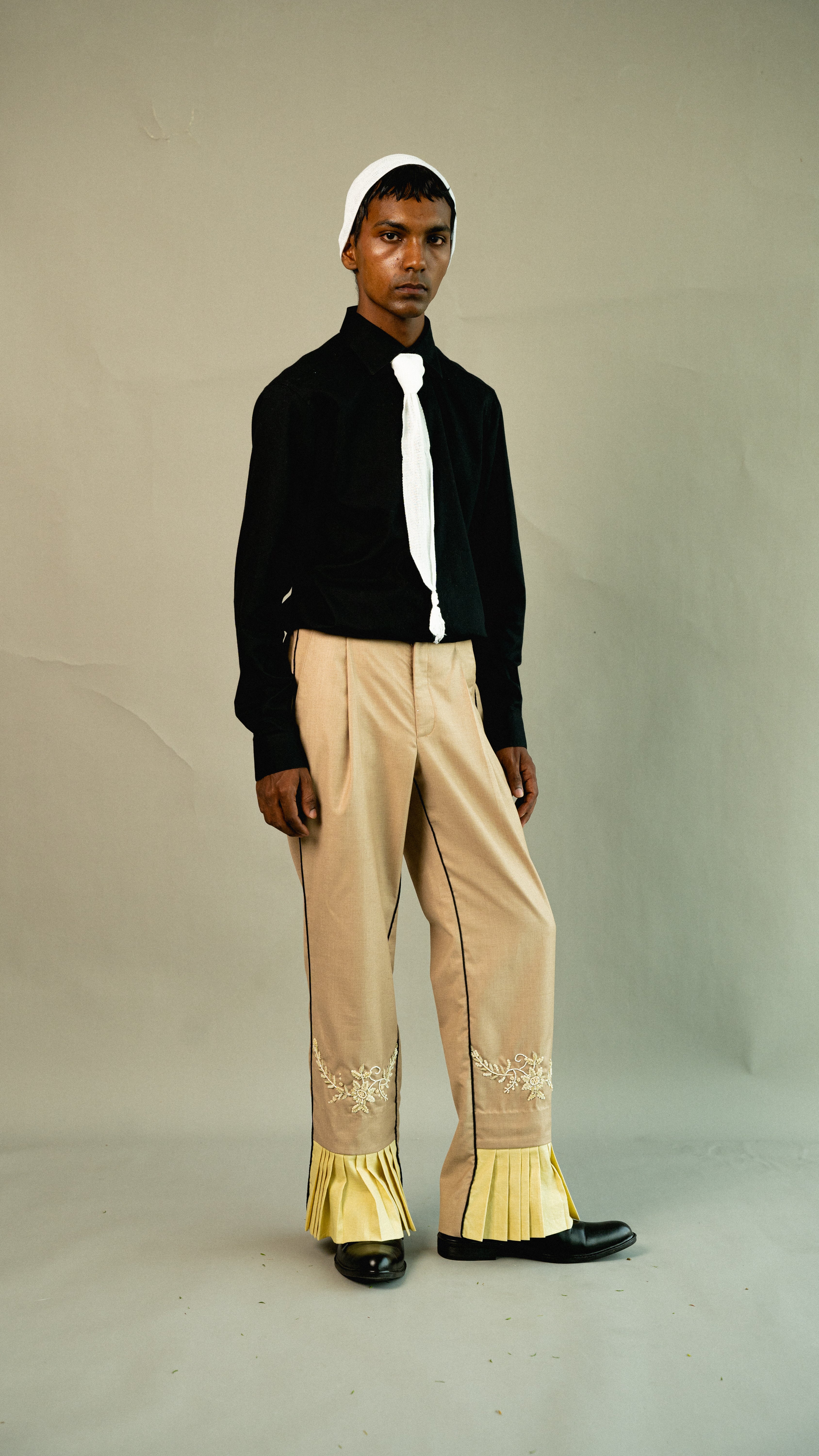 Man wearing a black sweater, beige pants with gold accents, and a white headscarf against a plain background