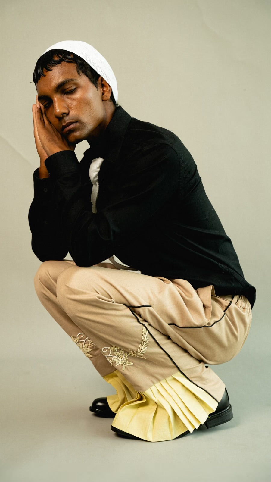 Man wearing a black sweater and beige pants with yellow accents, sitting on a light gray background.