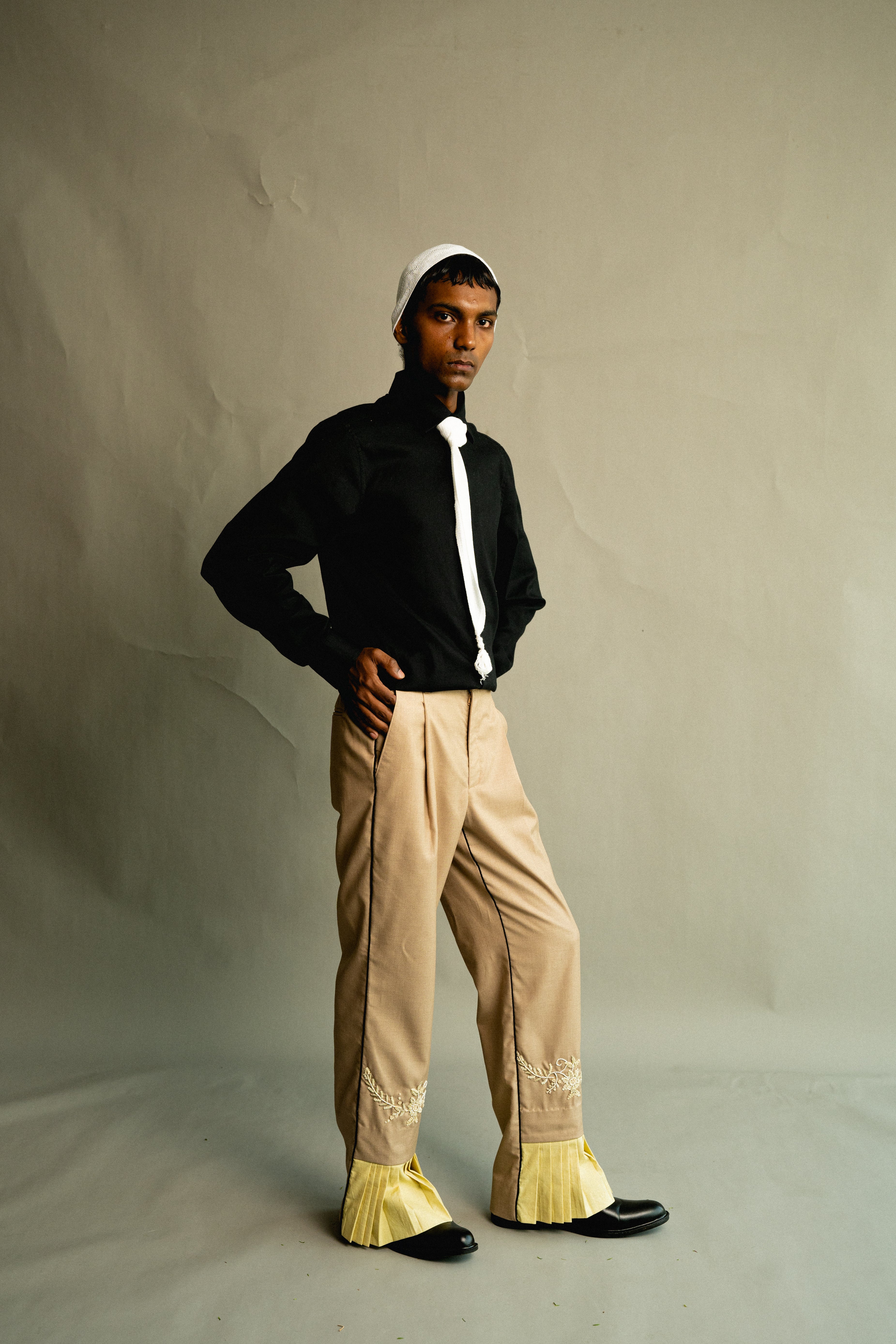 Person wearing a black shirt, beige pants with yellow accents, and a white cap against a plain background