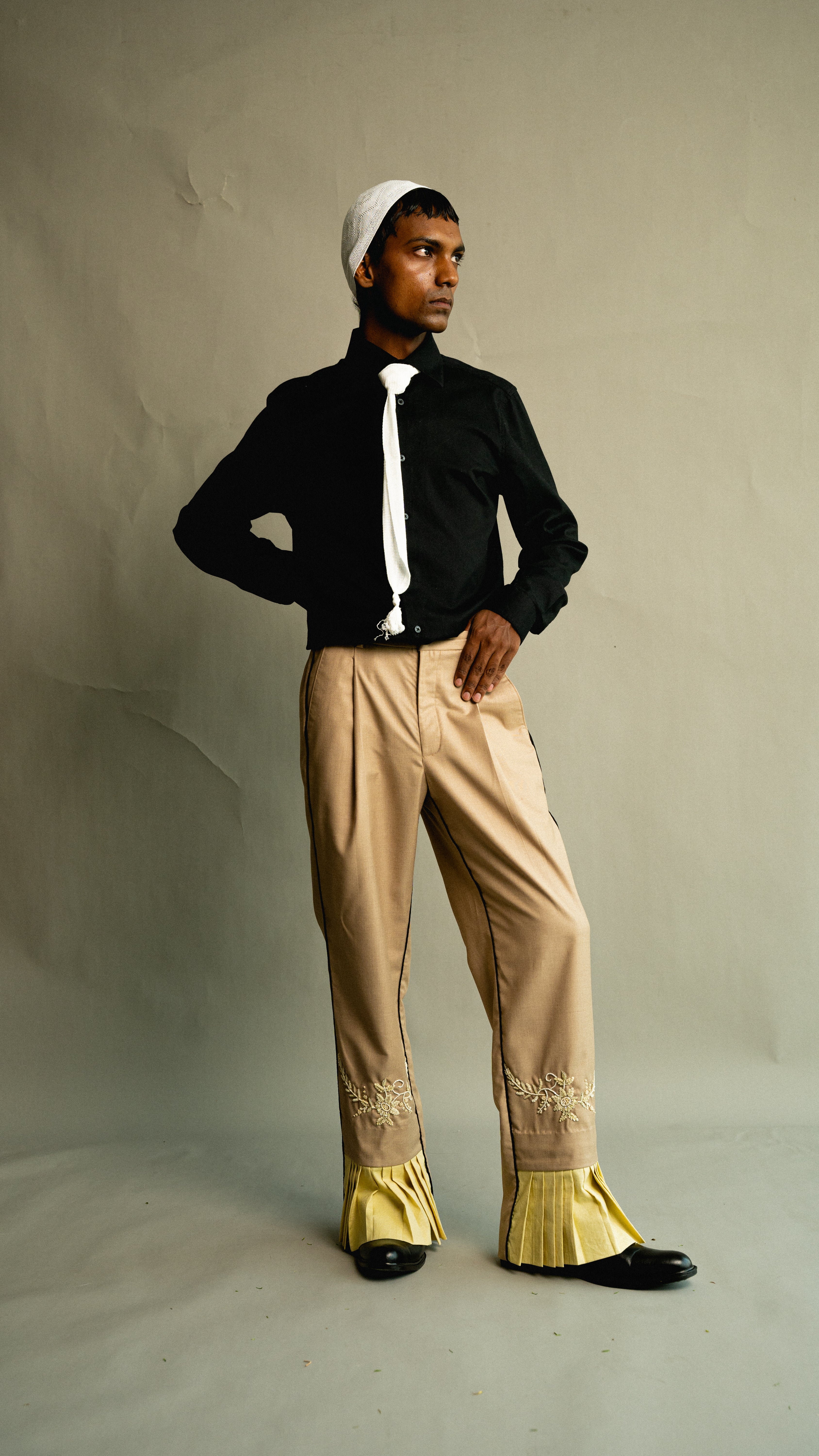Person wearing a black shirt, beige pants with gold trim, and a white headscarf against a plain background.
