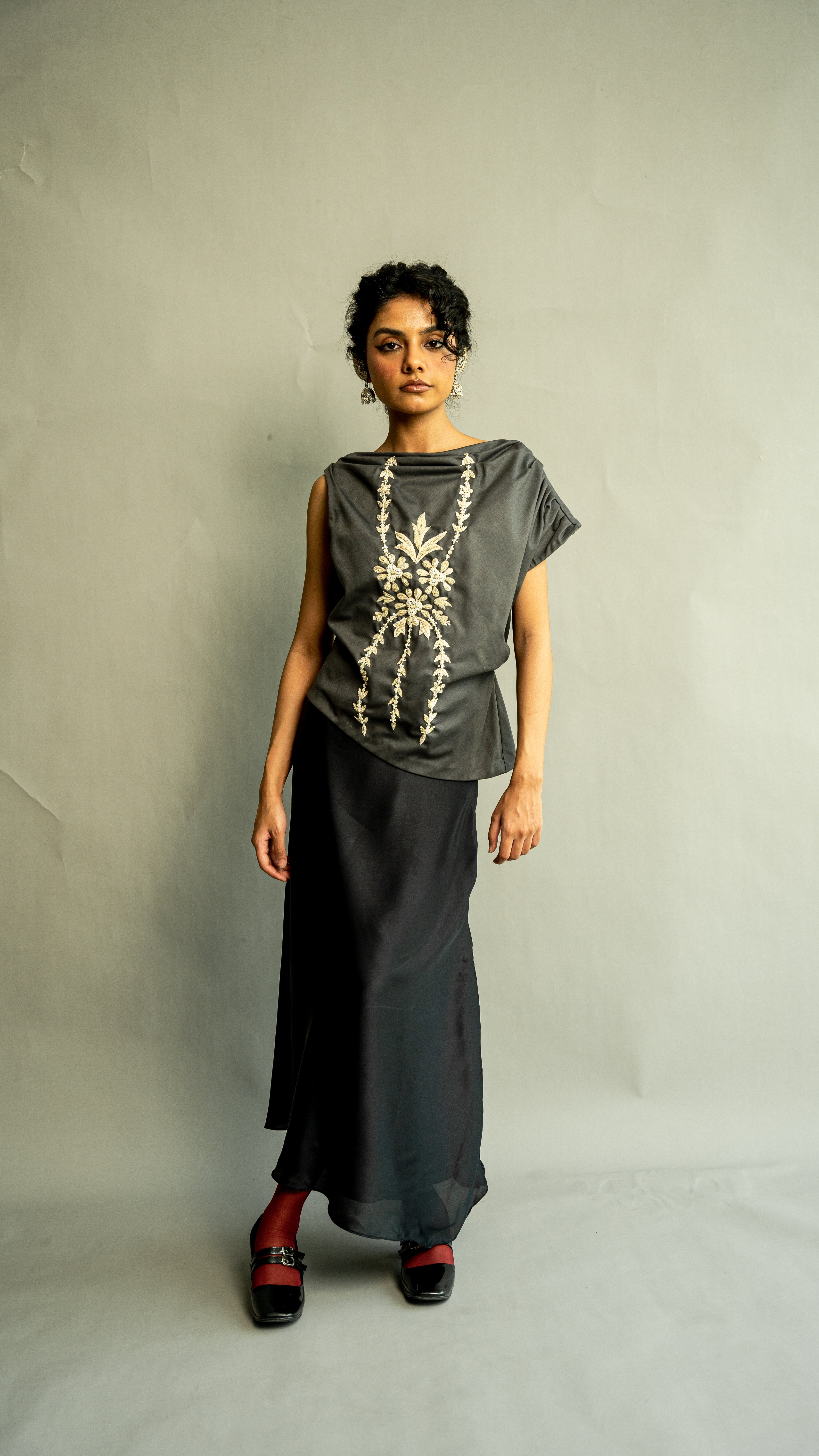 Person wearing a dark gray top with floral embroidery and a long black skirt on a plain background