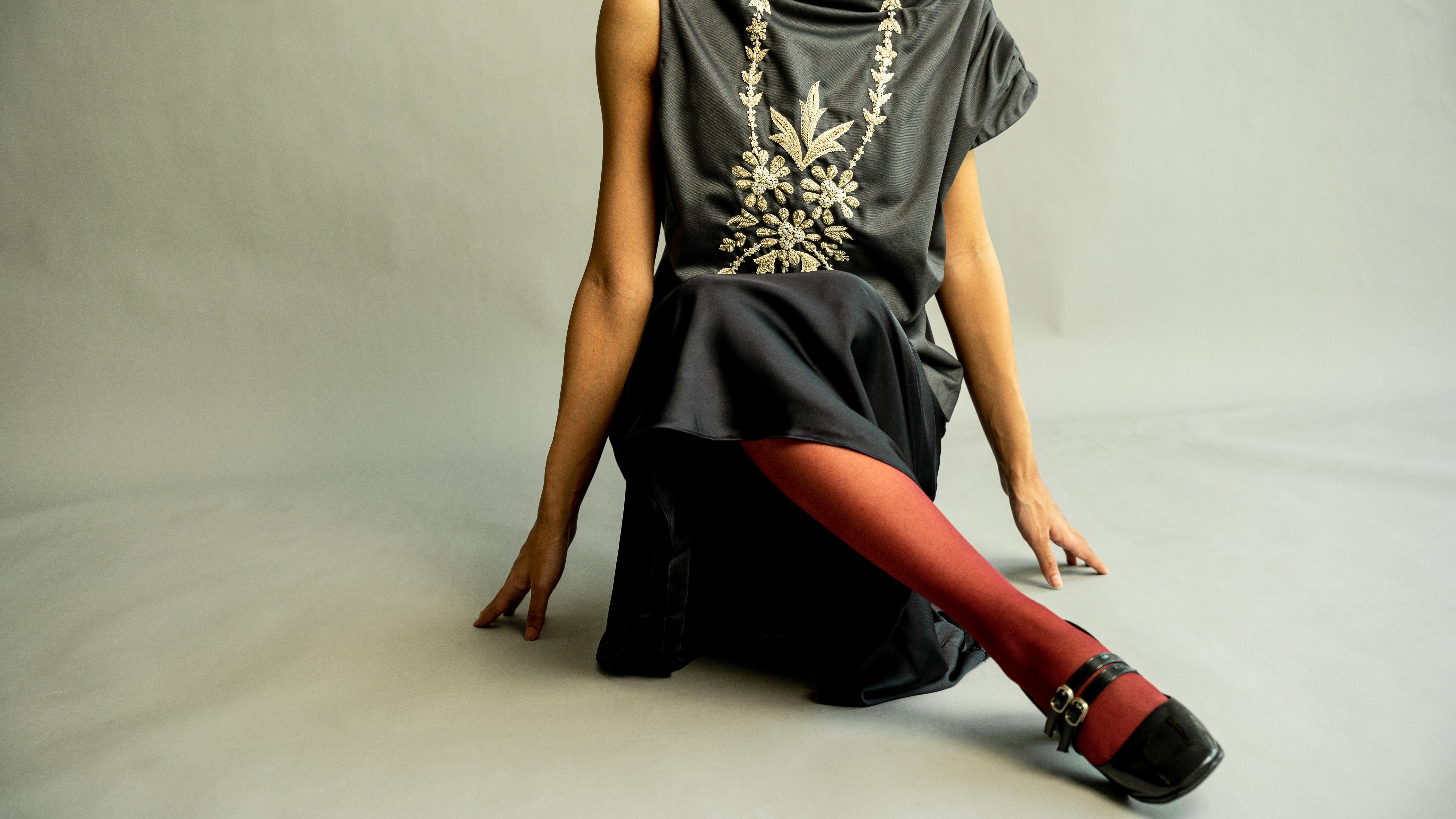 Person wearing a gray dress with floral embroidery, red tights, and black shoes on a neutral background