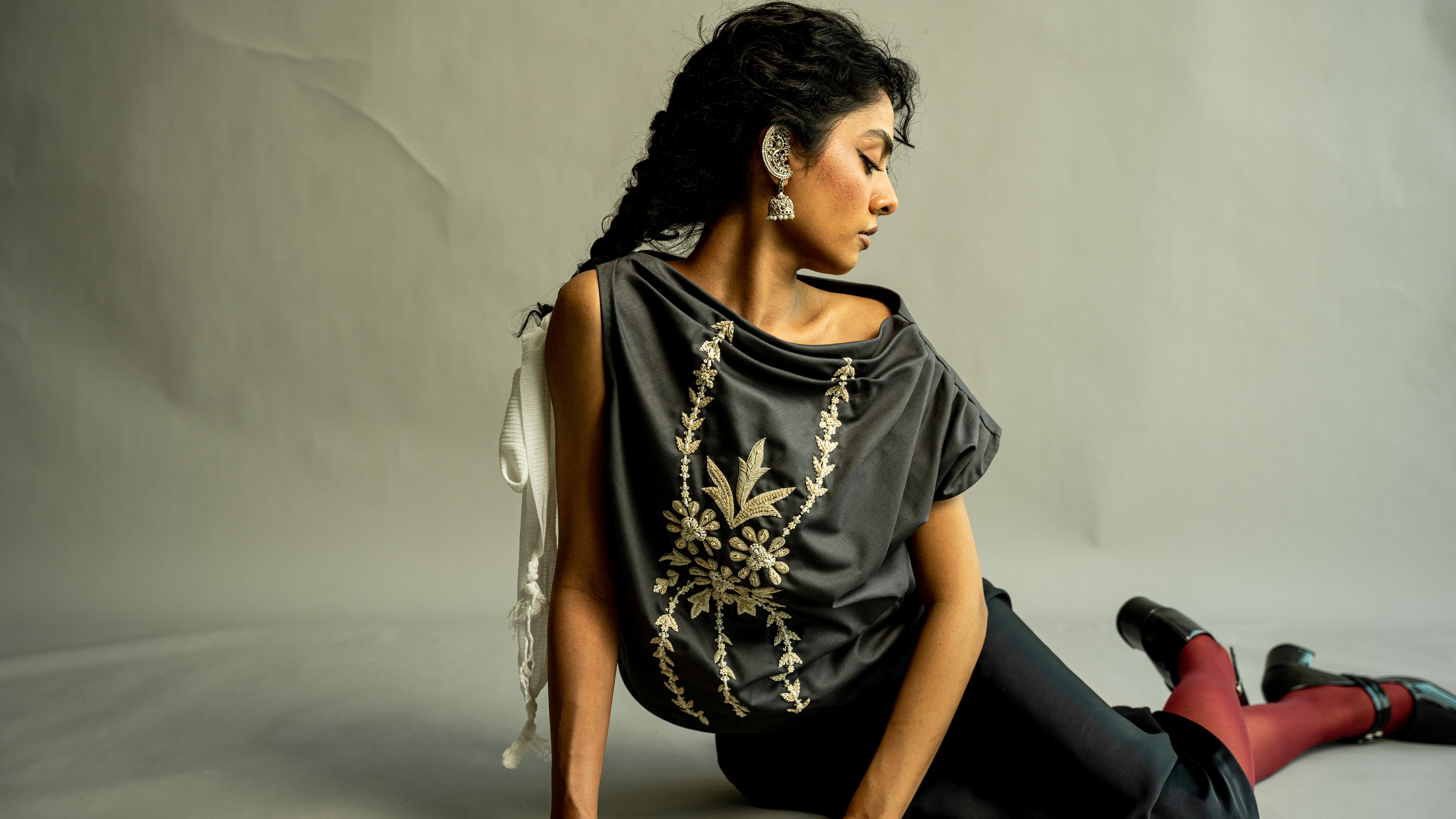 Woman sitting on a white surface wearing a dark gray top with floral embroidery and red pants.