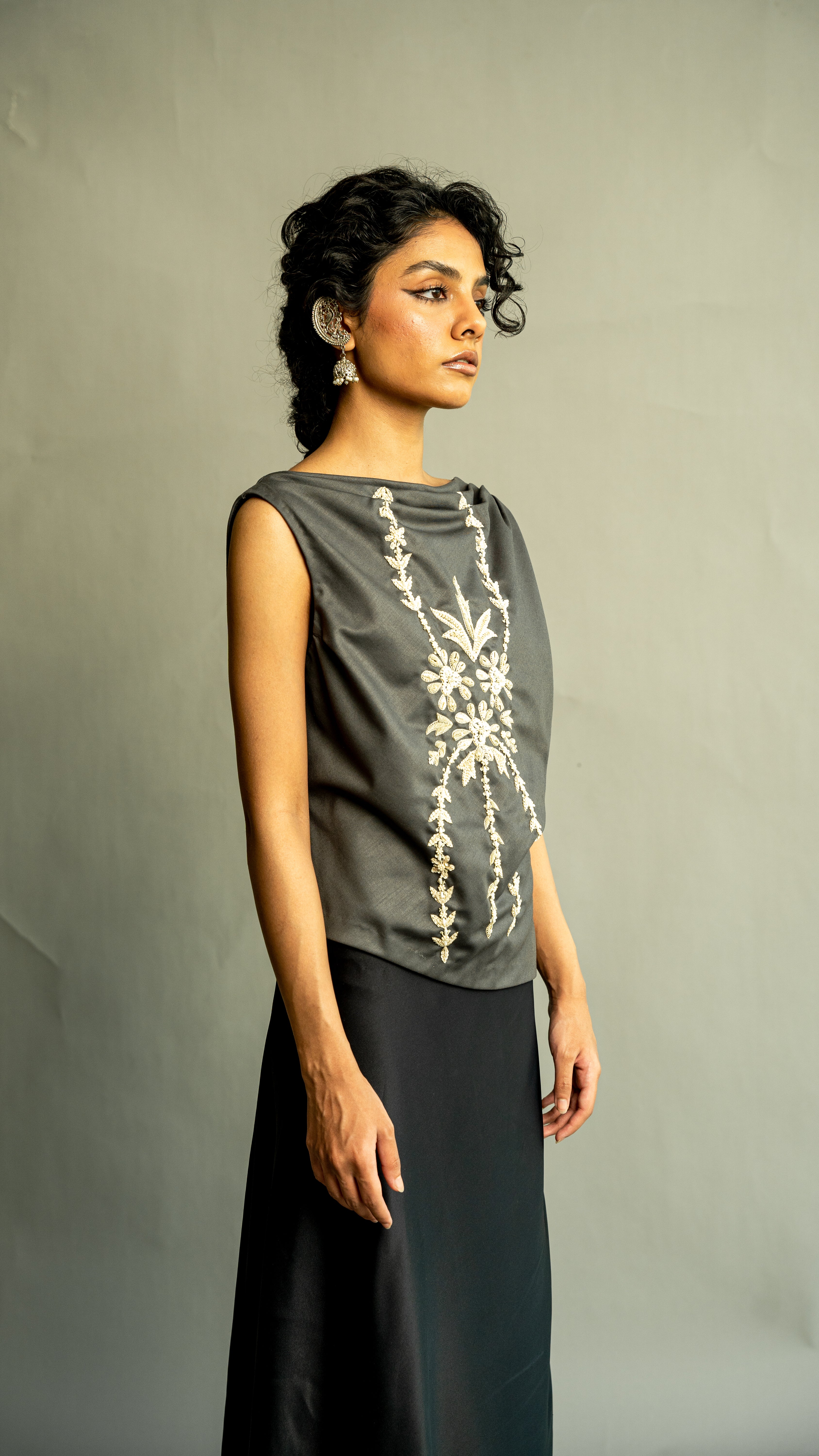 Woman wearing a gray sleeveless top with floral embroidery and black pants against a plain background