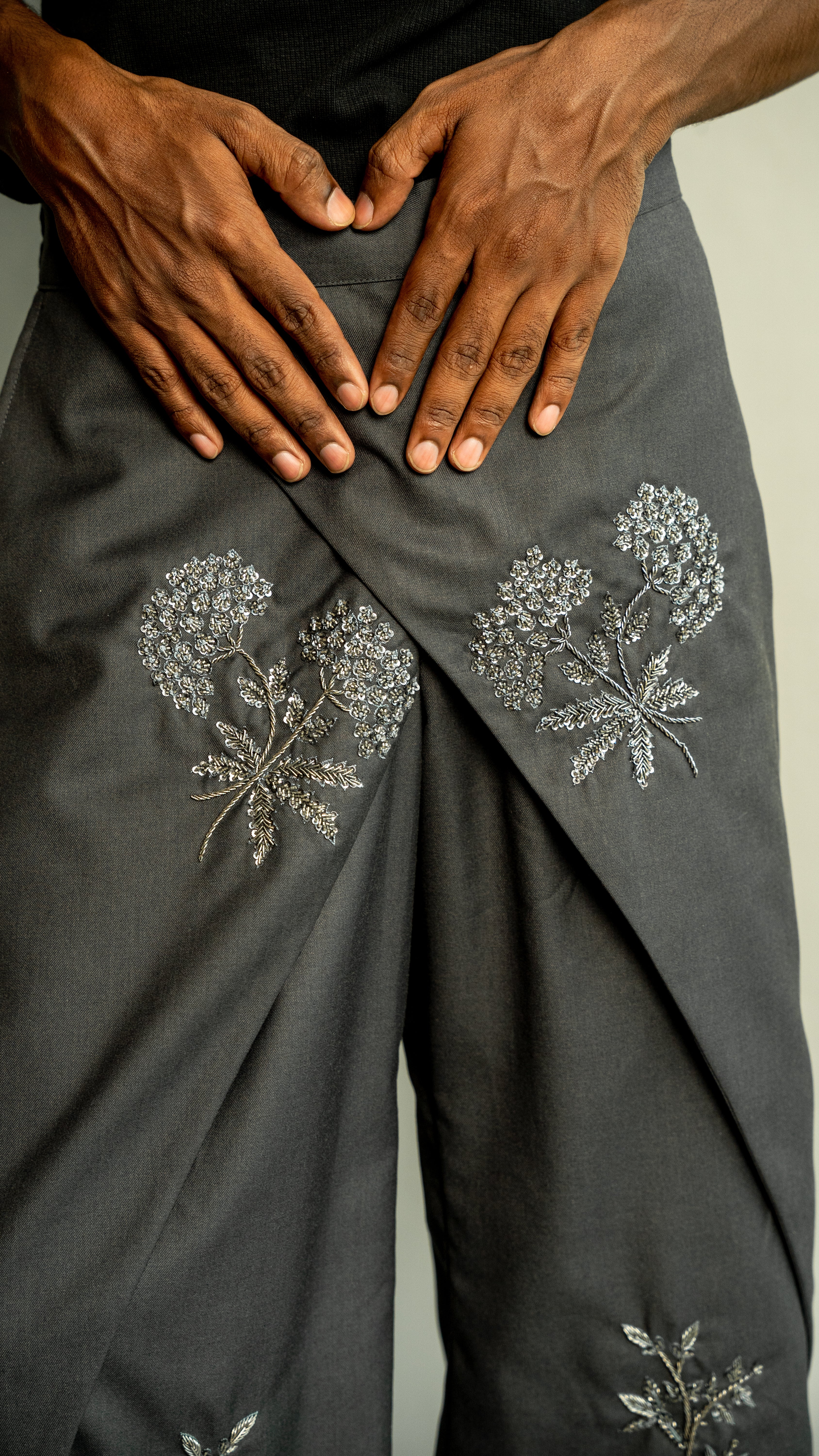 Dark gray pants with floral embroidery held by hands against a neutral background