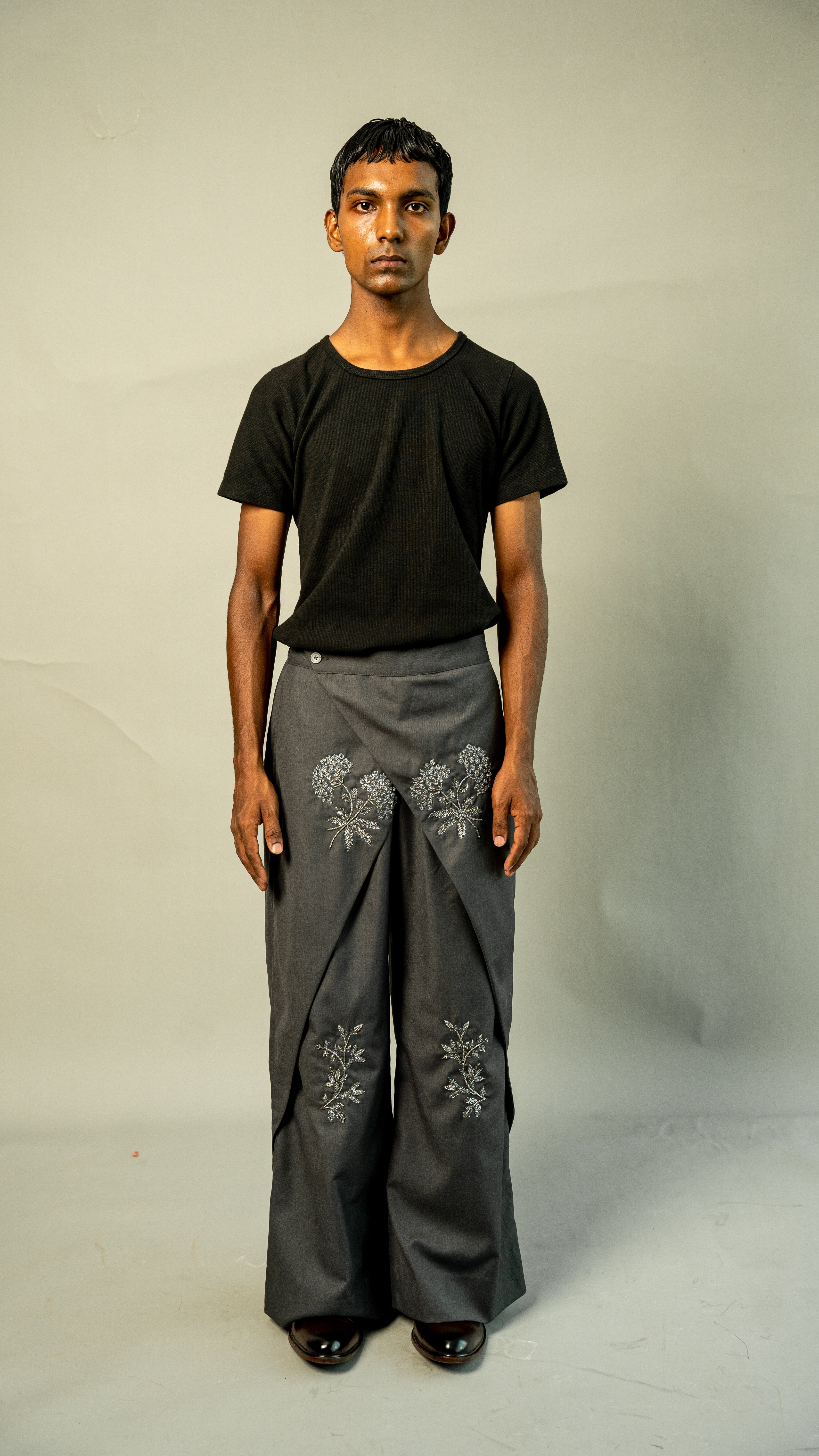 Man wearing a black t-shirt and gray pants with floral embroidery on a plain background